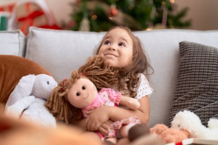 Photo for Adorable hispanic girl holding doll sitting on sofa by christmas tree at home - Royalty Free Image