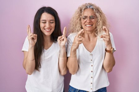 Photo for Mother and daughter standing together over pink background gesturing finger crossed smiling with hope and eyes closed. luck and superstitious concept. - Royalty Free Image