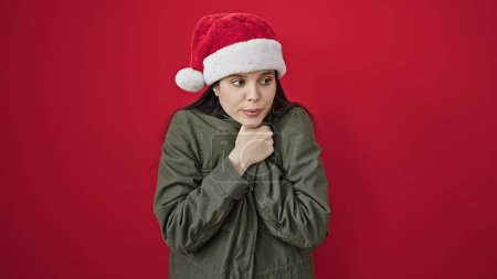 Foto de Joven mujer hispana hermosa usando sombrero de Navidad sufriendo por frío sobre fondo rojo aislado - Imagen libre de derechos
