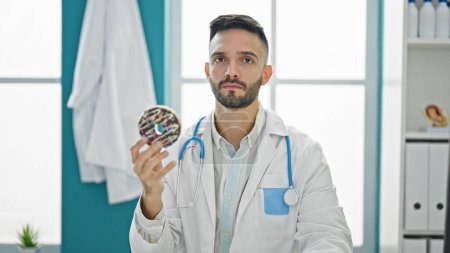 Foto de Joven médico hispano sentado en la mesa con expresión seria sosteniendo donut en la clínica - Imagen libre de derechos