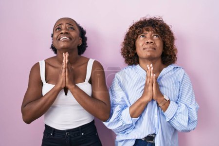 Téléchargez les photos : Deux femmes africaines debout sur fond rose suppliant et priant avec les mains ainsi que l'expression d'espoir sur le visage très émotionnel et inquiet. mendier. - en image libre de droit