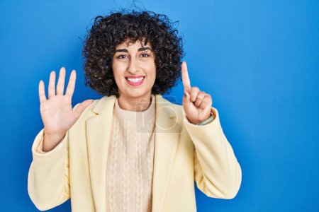 Téléchargez les photos : Jeune femme brune avec les cheveux bouclés debout sur fond bleu montrant et pointant vers le haut avec les doigts numéro six tout en souriant confiant et heureux. - en image libre de droit