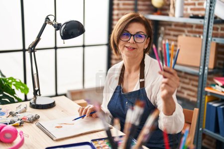 Photo for Middle age woman artist drawing on notebook holding pencils at art studio - Royalty Free Image
