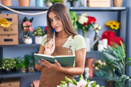Téléchargez les photos : Young beautiful hispanic woman florist reading notebook with doubt expression at flower shop - en image libre de droit
