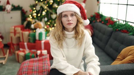 Foto de Mujer rubia joven sentada en el sofá por el árbol de Navidad con expresión relajada en casa - Imagen libre de derechos