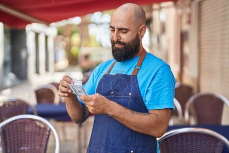 Téléchargez les photos : Jeune homme chauve serveur souriant confiant compter les dollars à la terrasse du café - en image libre de droit