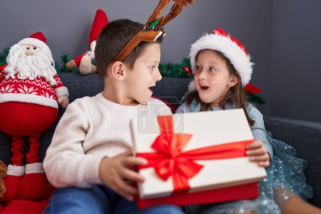 Téléchargez les photos : Adorable garçon et fille déballer cadeau de Noël assis sur le canapé à la maison - en image libre de droit