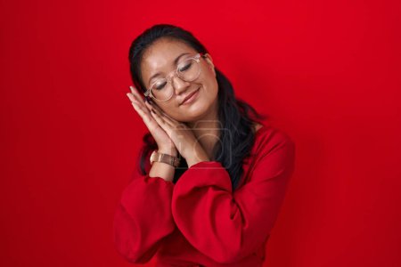 Foto de Asiática joven mujer de pie sobre rojo fondo durmiendo cansado soñando y posando con las manos juntas mientras sonríe con los ojos cerrados. - Imagen libre de derechos