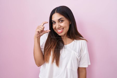 Téléchargez les photos : Jeune femme arabe debout sur fond rose geste souriant et confiant avec la main faisant signe de petite taille avec les doigts regardant et la caméra. concept de mesure. - en image libre de droit