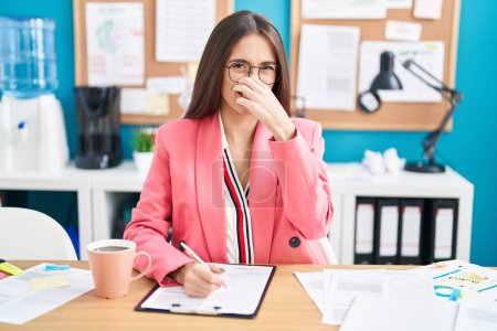 Photo for Young hispanic woman working at the office wearing glasses smelling something stinky and disgusting, intolerable smell, holding breath with fingers on nose. bad smell - Royalty Free Image