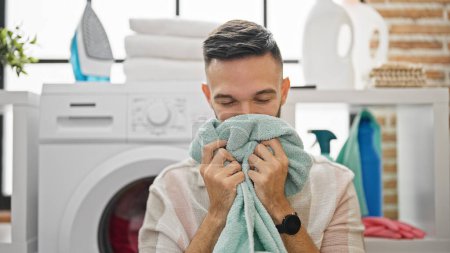 Photo for Young hispanic man smelling clean towel smiling at laundry room - Royalty Free Image