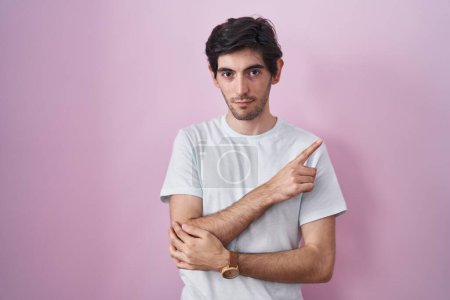 Photo for Young hispanic man standing over pink background pointing with hand finger to the side showing advertisement, serious and calm face - Royalty Free Image