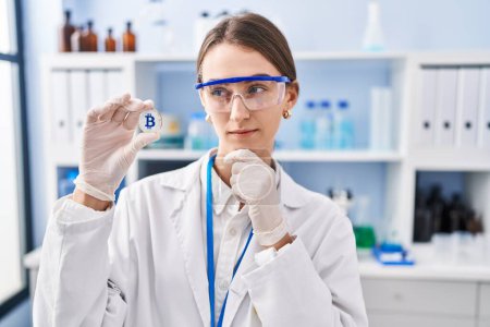 Photo for Young caucasian woman working at scientist laboratory holding bitcoin serious face thinking about question with hand on chin, thoughtful about confusing idea - Royalty Free Image