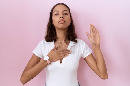 Téléchargez les photos : Jeune femme hispanique portant un t-shirt blanc décontracté jurant avec la main sur la poitrine et la paume ouverte, faisant un serment de fidélité - en image libre de droit