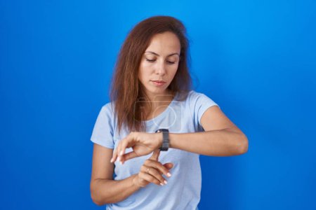 Téléchargez les photos : Femme brune debout sur fond bleu vérifier l'heure sur montre-bracelet, détendu et confiant - en image libre de droit