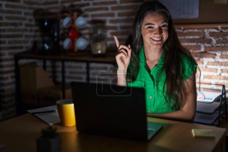 Téléchargez les photos : Jeune adolescente travaillant au bureau la nuit avec un grand sourire sur le visage, pointant la main et le doigt vers le côté en regardant la caméra. - en image libre de droit