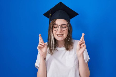 Photo for Blonde caucasian woman wearing graduation cap gesturing finger crossed smiling with hope and eyes closed. luck and superstitious concept. - Royalty Free Image
