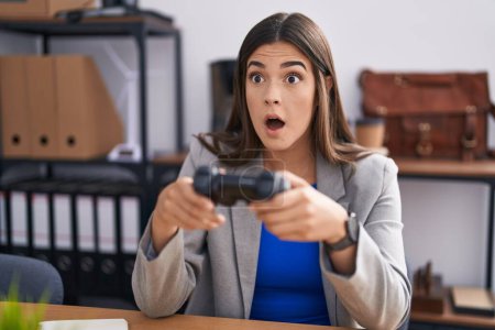 Téléchargez les photos : Femme hispanique travaillant au bureau jouant à des jeux vidéo effrayée et choquée par la surprise et l'expression étonnante, la peur et le visage excité. - en image libre de droit
