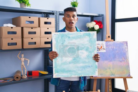 Photo for Young hispanic man holding canvas at artist studio afraid and shocked with surprise and amazed expression, fear and excited face. - Royalty Free Image