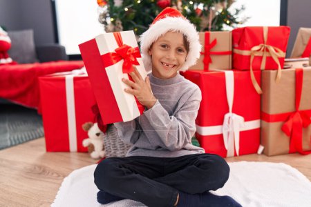 Foto de Adorable niño hispano sosteniendo regalo de Navidad sentado en el suelo en casa - Imagen libre de derechos