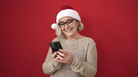 Téléchargez les photos : Femme d'âge moyen avec des cheveux gris portant un chapeau de Noël en utilisant un smartphone sur fond rouge isolé - en image libre de droit