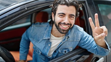 Photo for Young hispanic man smiling confident sitting on car doing victory gesture at street - Royalty Free Image