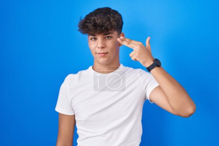 Foto de Adolescente hispano de pie sobre fondo azul disparando y suicidándose señalando la mano y los dedos a la cabeza como arma, gesto suicida. - Imagen libre de derechos
