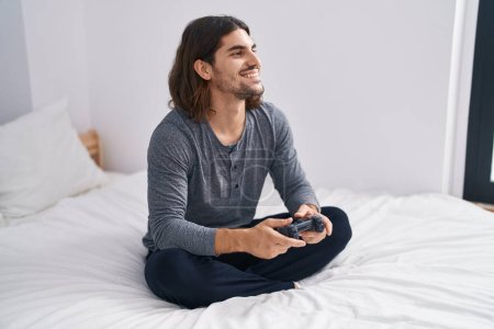 Photo for Young hispanic man playing video game sitting on bed at bedroom - Royalty Free Image