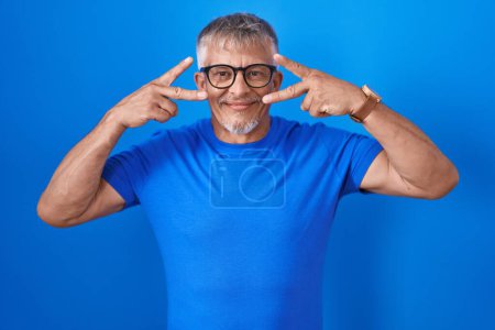 Téléchargez les photos : Homme hispanique avec les cheveux gris debout sur fond bleu faisant symbole de paix avec les doigts sur le visage, souriant joyeux montrant la victoire - en image libre de droit