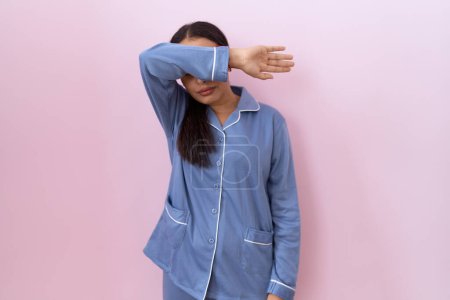 Foto de Mujer árabe joven con pijama azul cubriendo los ojos con el brazo, luciendo seria y triste. concepto de ocultamiento y rechazo sin visión - Imagen libre de derechos