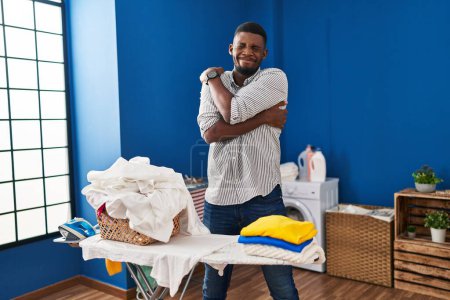 Photo for African american man ironing clothes at home hugging oneself happy and positive, smiling confident. self love and self care - Royalty Free Image