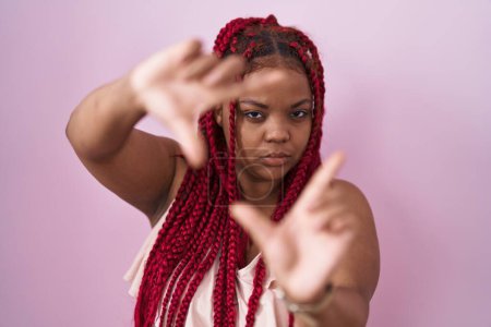 Foto de Mujer afroamericana con cabello trenzado de pie sobre fondo rosa haciendo marco usando las manos palmas y los dedos, perspectiva de la cámara - Imagen libre de derechos