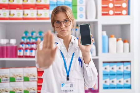Téléchargez les photos : Jeune femme caucasienne travaillant à la pharmacie pharmacie montrant écran de smartphone avec la main ouverte faisant stop signe avec expression sérieuse et confiante, geste de défense - en image libre de droit
