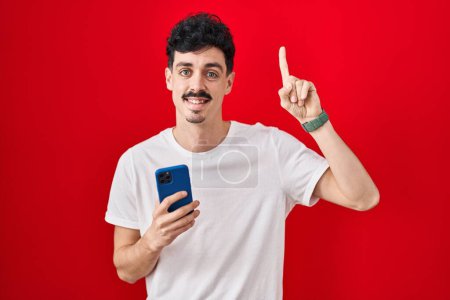 Téléchargez les photos : Homme hispanique utilisant smartphone sur fond rouge souriant étonné et surpris et pointant vers le haut avec les doigts et les bras levés. - en image libre de droit