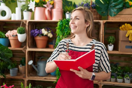 Téléchargez les photos : Jeune femme blonde fleuriste souriant écriture confiante sur ordinateur portable à la boutique de fleurs - en image libre de droit