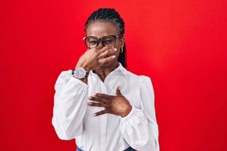 Téléchargez les photos : Femme africaine avec des tresses debout sur fond rouge sentant quelque chose de puant et dégoûtant, odeur intolérable, retenant souffle avec les doigts sur le nez. mauvaise odeur - en image libre de droit
