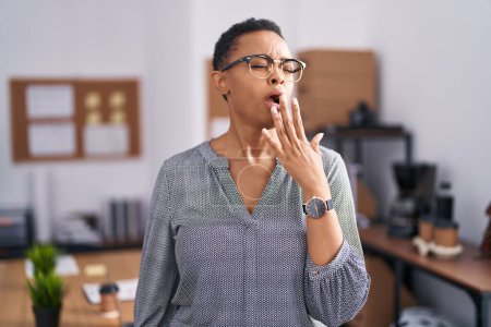 Foto de Mujer afroamericana trabajando en la oficina con gafas aburridas bostezando cansada cubriendo la boca con la mano. inquietud y somnolencia. - Imagen libre de derechos