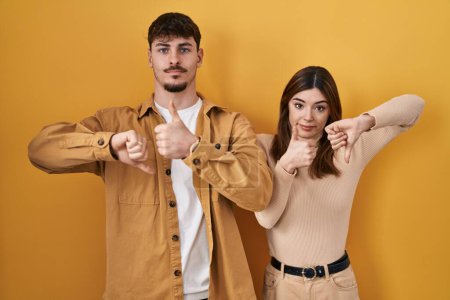 Photo for Young hispanic couple standing over yellow background doing thumbs up and down, disagreement and agreement expression. crazy conflict - Royalty Free Image