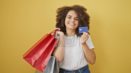 Foto de Joven mujer hispana hermosa sosteniendo bolsas de compras y tarjeta de crédito sonriendo sobre fondo amarillo aislado - Imagen libre de derechos