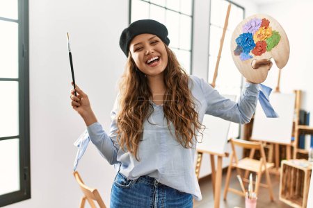 Téléchargez les photos : Jeune belle artiste hispanique souriante confiante tenant pinceau et palette au studio d'art - en image libre de droit