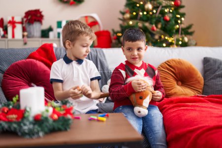 Foto de Dos niños jugando con juguetes sentados en el sofá junto al árbol de Navidad en casa - Imagen libre de derechos