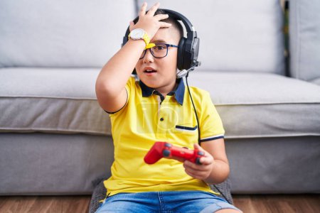 Téléchargez les photos : Jeune enfant hispanique jouant contrôleur de tenue de jeu vidéo portant des écouteurs surpris avec la main sur la tête pour erreur, rappelez-vous erreur. oublié, mauvais concept de mémoire. - en image libre de droit