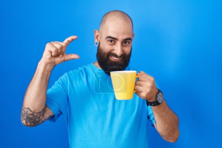 Téléchargez les photos : Jeune homme hispanique avec barbe et tatouages boire une tasse de café geste souriant et confiant avec la main faisant signe de petite taille avec les doigts regardant et la caméra. concept de mesure. - en image libre de droit