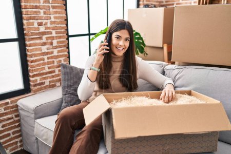 Photo for Young beautiful hispanic woman talking on smartphone unpacking cardboard box at new home - Royalty Free Image