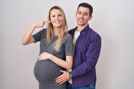 Téléchargez les photos : Jeune couple s'attendant à un bébé debout sur fond blanc souriant gai montrant et pointant avec les doigts dents et la bouche. concept de santé dentaire. - en image libre de droit
