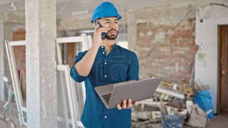 Photo for Young hispanic man architect wearing hardhat using laptop speaking on the phone at construction site - Royalty Free Image