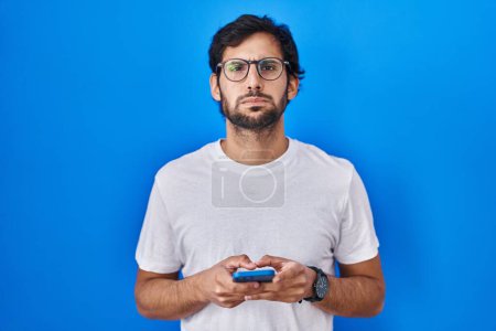 Photo for Handsome latin man using smartphone typing message puffing cheeks with funny face. mouth inflated with air, catching air. - Royalty Free Image