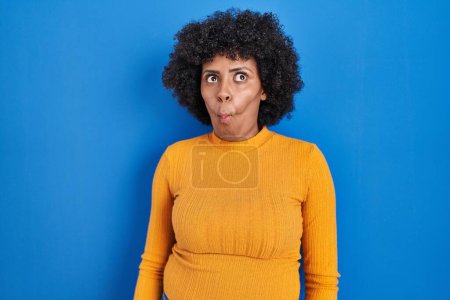 Photo for Black woman with curly hair standing over blue background making fish face with lips, crazy and comical gesture. funny expression. - Royalty Free Image