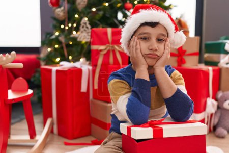 Photo for Adorable hispanic boy holding christmas gift sitting on floor with unhappy expression at home - Royalty Free Image