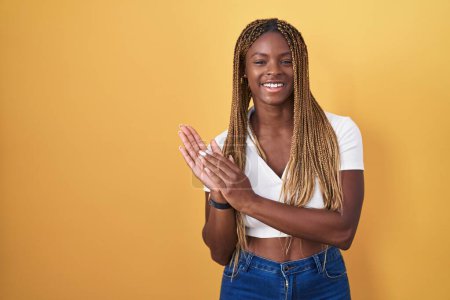 Foto de Mujer afroamericana con el pelo trenzado de pie sobre el fondo amarillo aplaudiendo y aplaudiendo felices y alegres, sonriendo orgullosas manos juntas - Imagen libre de derechos